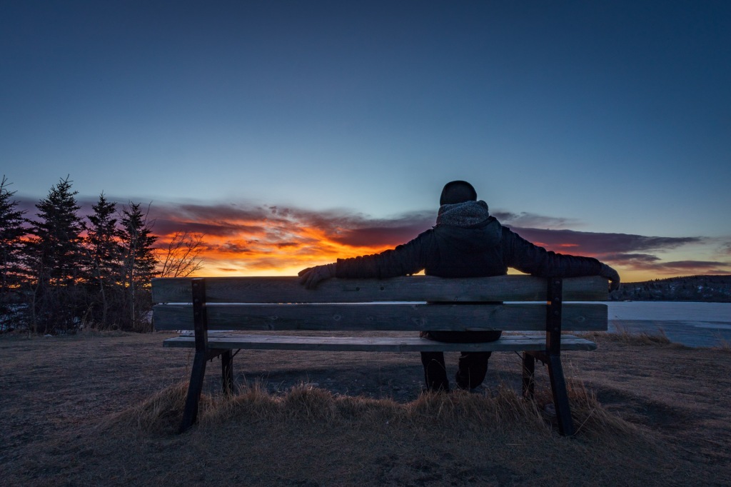 A man sitting on a park bench is silhouetted by a fiery cloud during sunrise at Ghost Lake, east of Cochrane, Alberta, Canada.  Photographed on March 20, 2021.